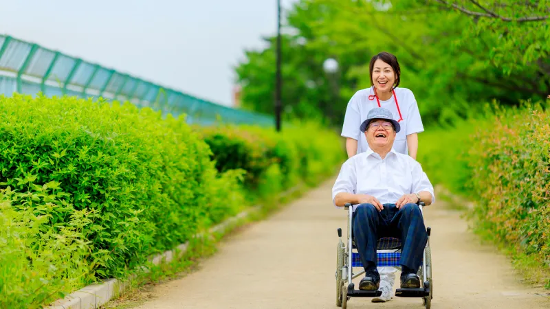 緑の中、おじいさんの乗った車椅子を押す白衣姿の女性看護師